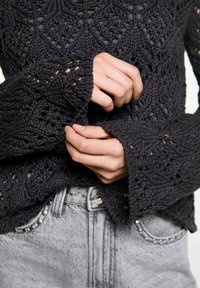 Dunkelgrauer Strickpullover mit einem strukturierten, gemusterten Design und ausladenden Ärmeln. Getragen über hellgrauen Jeans mit metallischen Akzenten.
