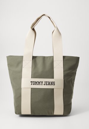 Bolso tote de lona verde con asas de tela crema, que presenta "TOMMY JEANS" impreso en la parte frontal. Forma cuadrada y textura duradera.