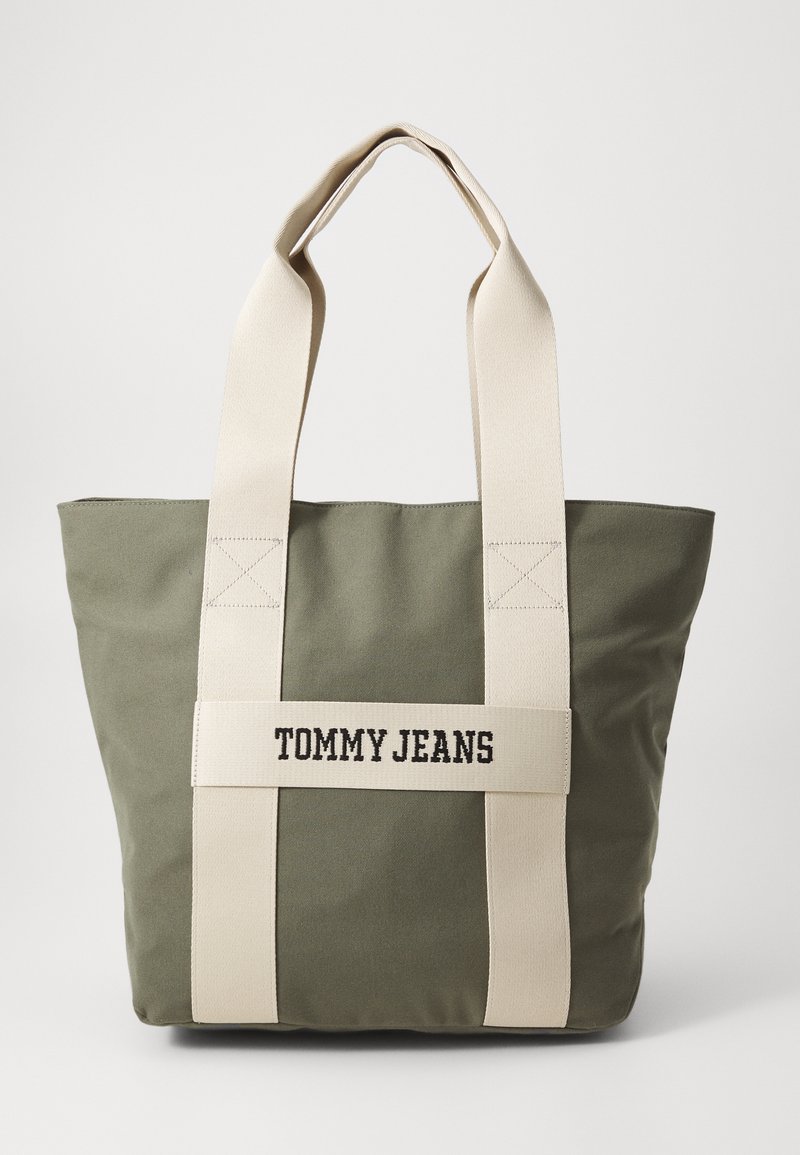 Green canvas tote bag with cream fabric straps, featuring "TOMMY JEANS" printed on the front. Square shape and durable texture.