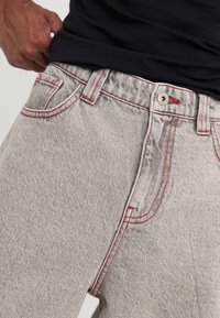 Jeans de mezclilla gris con costuras rojas, que cuentan con un diseño de cinco bolsillos, cierre de botón y textura visible en la tela.
