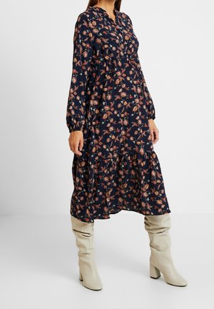 Kvinde iført en navyblå blomsterprintet langærmet midikjole kombineret med lysebeige slouchy hælede støvler, står foran en ensfarvet baggrund.