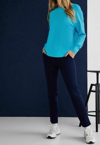 Turkos långärmad topp i kombination med marinblå byxor. Modellen bär vita sneakers. Omgivningen har mörka bakgrunder och en svart pall.