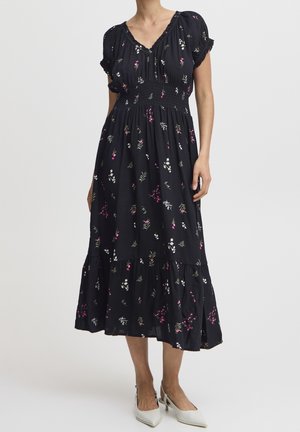 Vrouw draagt een zwarte midi-jurk met roze en witte bloemenprints, korte pofmouwen, een geplooide taille en witte pumps met puntige neus.