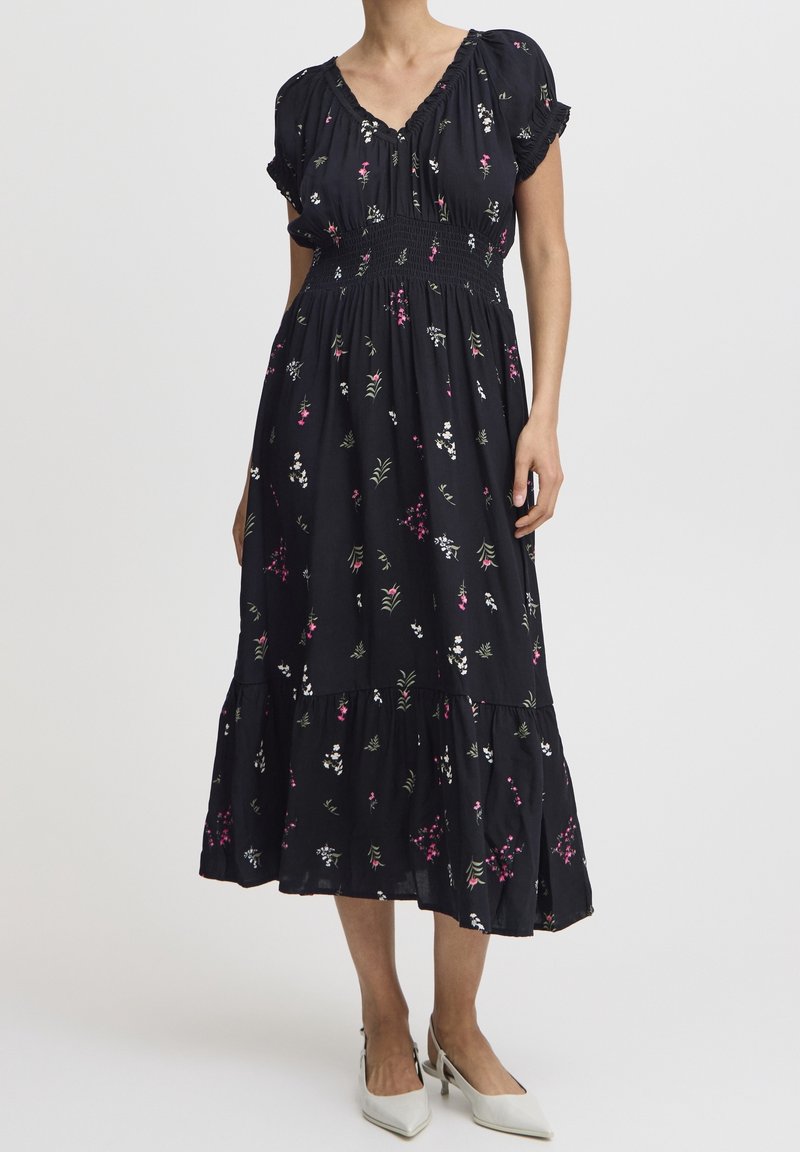 Femme portant une robe midi noire avec des imprimés floraux roses et blancs, des manches courtes bouffantes, une taille froncée et des talons blancs pointus.