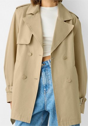 Trench-coat beige croisé sur un crop top blanc et un jean denim taille haute bleu clair sur une personne debout.