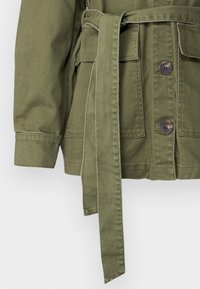 Olive green jacket z paskiem w talii, z czterema guzikami z przodu, dwiema kieszeniami i kontrastującymi szwami. Miękki, teksturowany materiał.