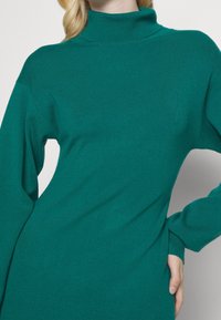 Pull col roulé bleu sarcelle avec une coupe décontractée, une texture côtelée, des manches longues et un col haut. Il comporte une poche latérale au niveau de la taille.