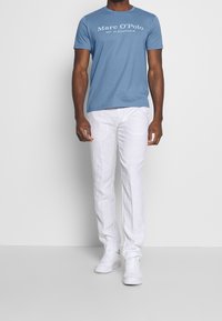 Camiseta de algodón azul con texto del logo en blanco, combinada con pantalones de lino blancos y zapatillas blancas, con cuello redondo y mangas cortas.