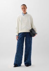 Weißer Pullover mit Schädel- und Kreuzbones-Design, kombiniert mit weit geschnittenen blauen Jeanshosenträgern und einer kleinen schwarzen Tasche. Glatte Textur.
