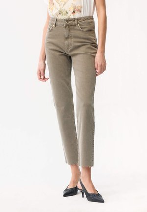 Kobieta ubrana w oliwkowe jeansy o prostych nogawkach i wysokim stanem z surowym finiszem oraz czarne sandały na niskim obcasie na palce, stojąca na białym tle.