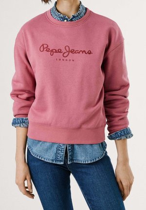 Osoba ubrana w różową bluzę Pepe Jeans London na podwiniętą koszulę jeansową i niebieskie dżinsy, pokazana od szyi do bioder.