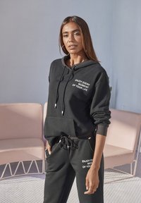 Schwarzer Hoodie und Jogginghose aus weichem Material, mit weißem Text "JEDEN MOMENT DEINES LEBENS GENIESSEN." Beinhaltet eine Fronttasche und einen Kordelzug.