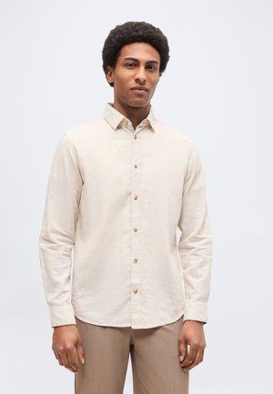 Homme aux cheveux courts et bouclés portant une chemise beige à boutons et un pantalon marron clair, debout devant un fond uni.