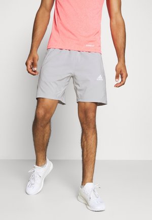 Mężczyzna ubrany w jasnoszorty Adidas, koralowo-różową koszulkę i białe trampki, stojący na jednolitym jasnym tle.