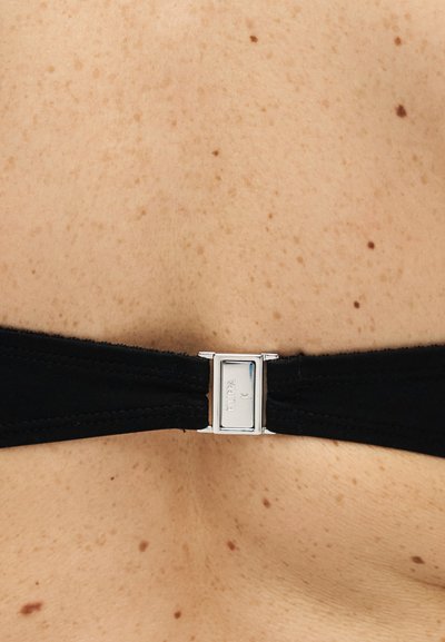 Κοντινό πλάνο σε μια πλάτη με φακίδες, με έναν μαύρο υφασμάτινο ιμάντα που στερεώνεται με μια ασημένια μεταλλική αγκράφα χαραγμένη με "FILIPPA K".