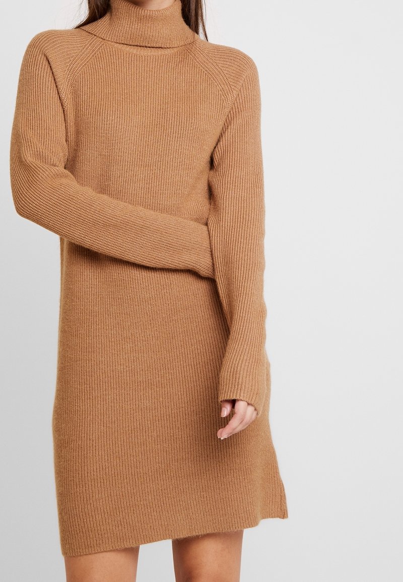 Robe en tricot couleur tan chaud, avec un col roulé haut, une texture côtelée et de longues manches avec des ouvertures pour les pouces pour un confort supplémentaire.