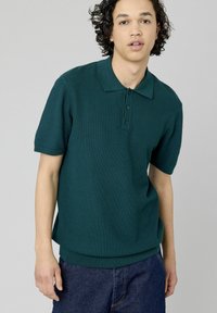 Polo en tricot vert foncé avec un col, patte de boutonnage à trois boutons, manches courtes et détails en côte au niveau de l'ourlet.