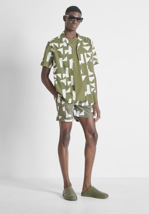 Jeune homme portant des lunettes de soleil et une chemise verte à manches courtes à motifs avec un short assorti et des chaussures slip-on sur un fond uni.