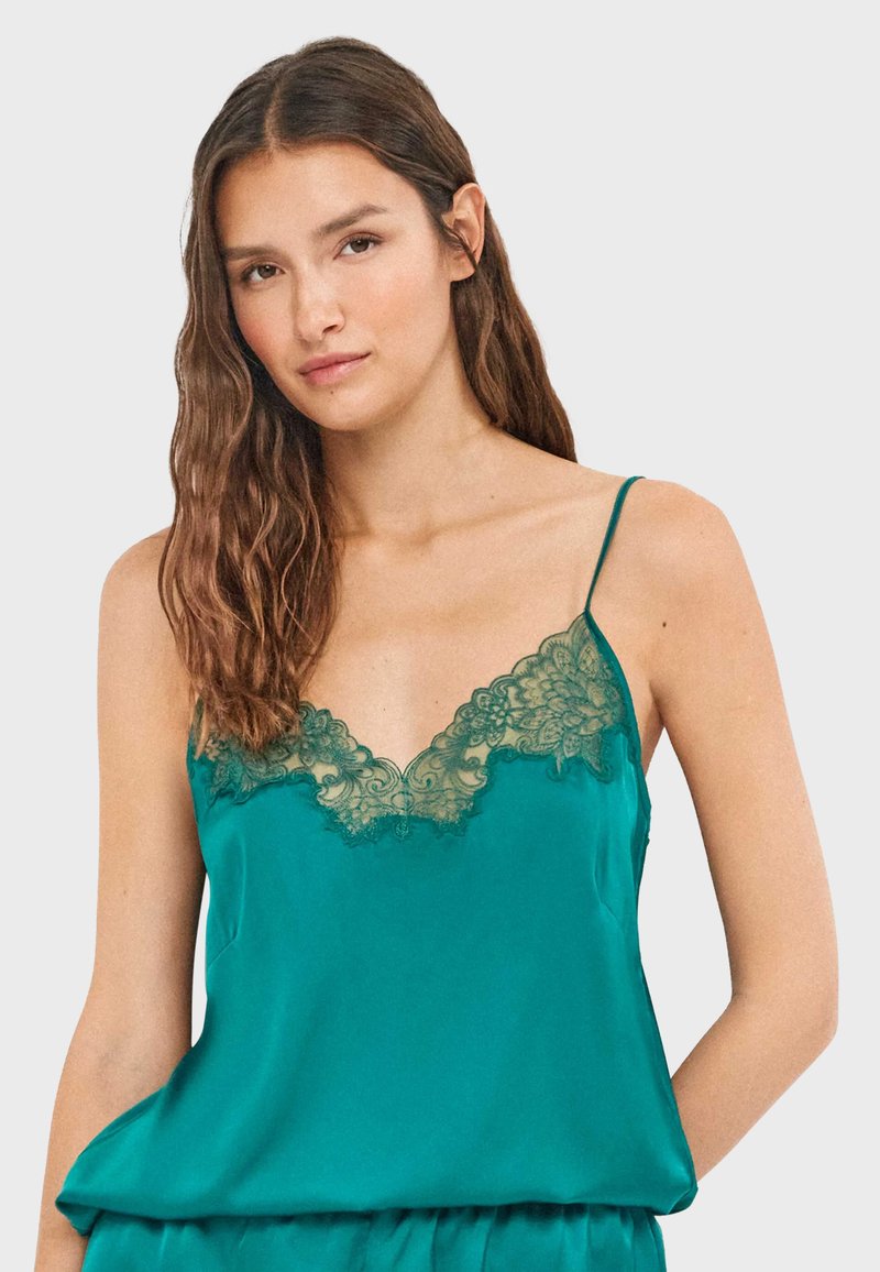 Jeune femme aux longs cheveux bruns portant un caraco en satin bleu sarcelle avec une bordure en dentelle verte et des bretelles fines, debout devant un fond uni.