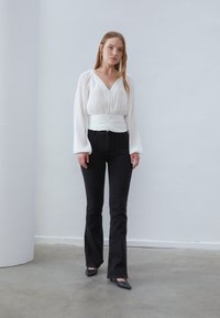 Hvid plisseret top med V-hals og indsnævret talje, parret med sorte flared jeans. Sorte spidse sko fuldender outfittet.