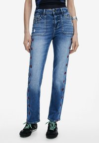 Sinine denimist teksad otsejoone lõikega, millel on punased lillemustrid külgedel, kulunud välimus ja viie tasku disain.