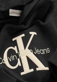 Felpa nera con logo "Calvin Klein Jeans" ricamato in crema. Caratterizzata da polsini e orlo a costine, tessuto morbido e cappuccio con cordini.