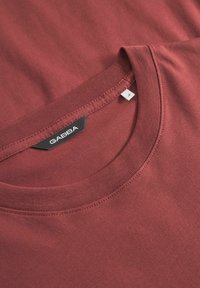 Bordeauxrød T-shirt i bomuld med rund halsudskæring, korte ærmer og en sort mærkat med teksten "GABBA" på kraven. Glat tekstur.