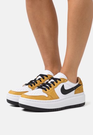 Jalat, joissa valkoiset ja sinapinkeltaiset Nike-tennarit mustalla Swoosh-logolla, mustilla nauhoilla ja paksuilla valkoisilla pohjilla yksivärisellä taustalla.