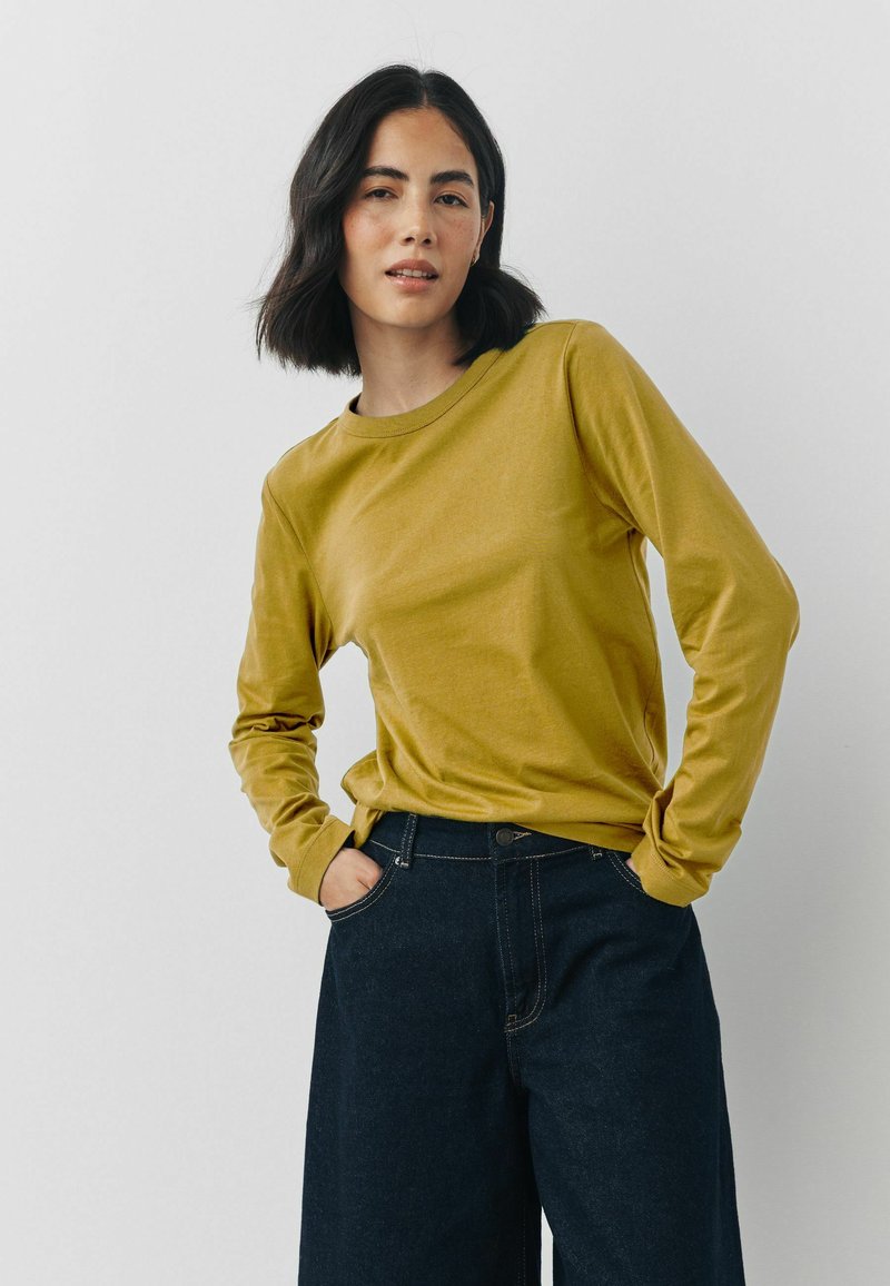 Langærmet, sennepsgul t-shirt lavet af blødt stof, med rund hals og afslappet pasform, paired med mørk denim bukser med bred ben.