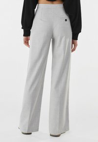 Kobieta nosząca jasnoszare, szerokie spodnie z wysokim stanem o delikatnym wzorze w kratkę i pojedynczej zapinanej na guzik tylnej kieszeni, w zestawieniu z czarną bluzką.