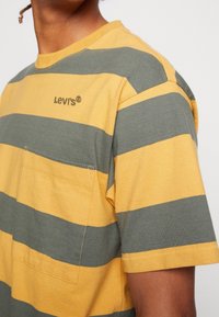 Tričko s krátkým rukávem, kulatým výstřihem a pruhy v hořčičně žluté a olivově zelené barvě z bavlny. Má přední kapsu a vyšitý logotyp značky.