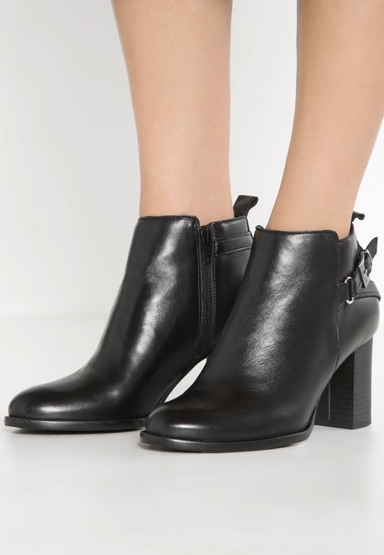 Bottines en cuir noir à talons blocs, avec fermeture éclair latérale et sangles décoratives à boucle, portées sur des jambes nues.