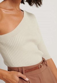 Top in maglia a coste color beige chiaro con un ampio scollo a spalle scoperte e maniche corte, abbinato a pantaloni marroni a vita alta e una cintura marrone.