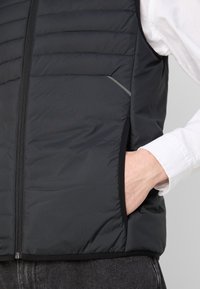 Homme portant un gilet matelassé noir avec la main dans la poche latérale, chemise blanche à manches longues et pantalon foncé, visible de la taille à la poitrine.