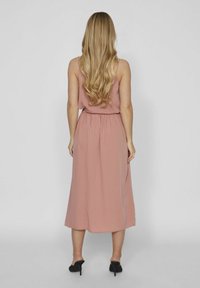 Vestido maxi sin mangas de color rosa con cintura elástica y falda fluida. Tela suave con un brillo sutil. Zapatos de tacón alto negros.
