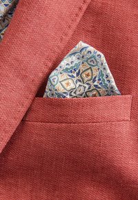 Rote strukturierte Blazer-Tasche mit einem gefalteten gemusterten Einstecktuch, das blaue, orange und grüne Blumenmotive auf einem hellen Hintergrund zeigt.