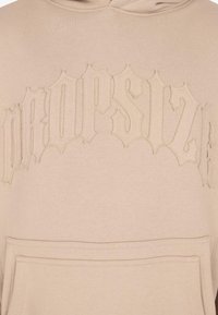 Un sweat à capuche beige avec du texte gothique brodé "PURPOSE" sur la poitrine et une poche kangourou à l'avant.