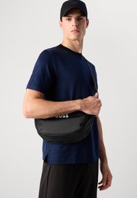 Saco cruzado preto com tecido texturizado, apresentando um logótipo branco. Usado sobre uma t-shirt listrada azul-marinho com decote redondo e calças escuras.