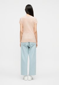 Γυναίκα με μακριά σκοτεινά μαλλιά, φοράει ανοιχτό ροζ κοντομάνικη μπλούζα, ανοιχτό μπλε τζιν με φαρδιά μπατζάκια και άσπρα αθλητικά, όρθια με την πλάτη στραμμένη.