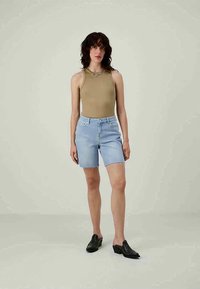 Light blue denim shorts with frayed hem; paired with a beige tank top. Model wears black slip-on shoes. Neutral background.