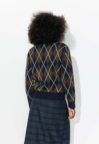 Pull en jacquard à motifs argyle marron, bleu marine et vert, avec un ourlet ajusté côtelé et des manches longues, associé à une jupe à carreaux dans des couleurs sombres.
