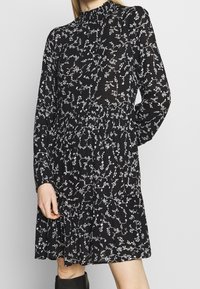 Robe noire avec un motif floral blanc, manches longues, col montant et taille froncée, confectionnée dans un tissu léger.