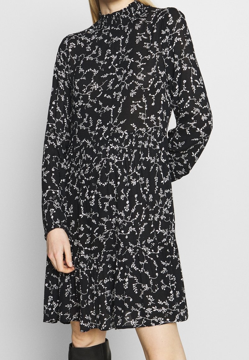 Robe noire avec un motif floral blanc, manches longues, col montant et taille froncée, confectionnée dans un tissu léger.