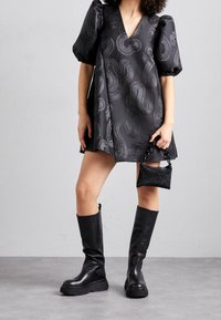 Schwarzes Kleid mit Puffärmeln, V-Ausschnitt und Wellenmuster. Accessoires sind eine kleine schwarze funkelnde Handtasche und kniehohe schwarze Stiefel.