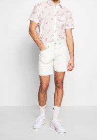 Camicia a maniche corte con bottoni color rosa chiaro con stampa di fenicotteri, abbinata a pantaloni corti in denim bianchi con orlo leggermente sfilacciato.