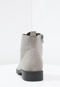 Stivaletto in suede grigio chiaro con cerniera sul retro, linguetta e suola nera visto da dietro su una superficie bianca.