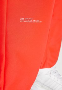 Röd sporttyg med vit text "Född från sport, bärs för stil, möjliggör självuttryck och komfort" ovanför vita sneakers.