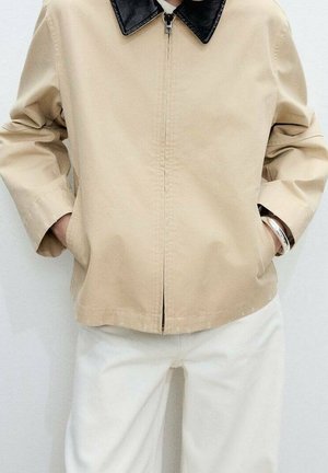 Veste beige avec col en cuir noir boutonnée, mains dans les poches, associée à un pantalon blanc et un bracelet argenté au poignet droit.