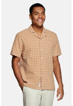 Lachende man met een korte mouw beige overhemd met wit geometrisch patroon en lichtgekleurde broek, staand met één hand in de zak.