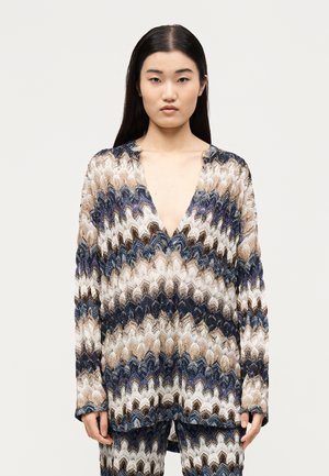 Kobieta z długimi ciemnymi włosami nosi luźny, wzorzysty sweter z dzianiny i pasujące spodnie w niebiesko-beżowo-białym wzorze jodełki.